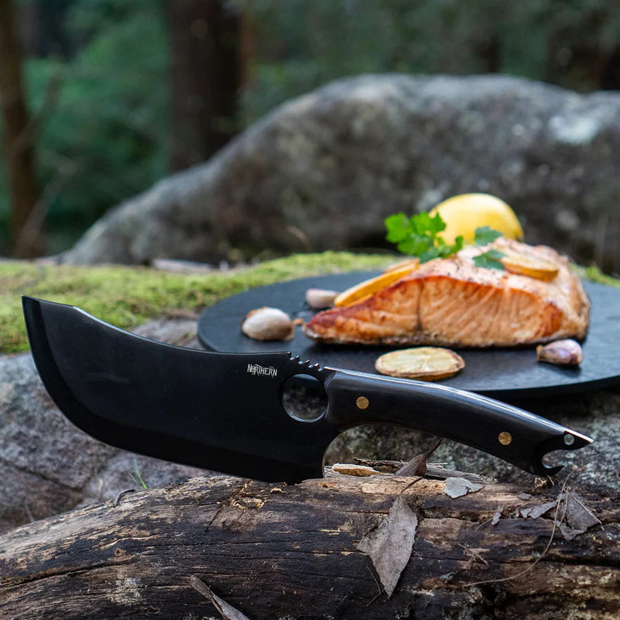 Black Viking knife placed beside grilled salmon on a forest log — showcasing elegance, precision, and outdoor utility.